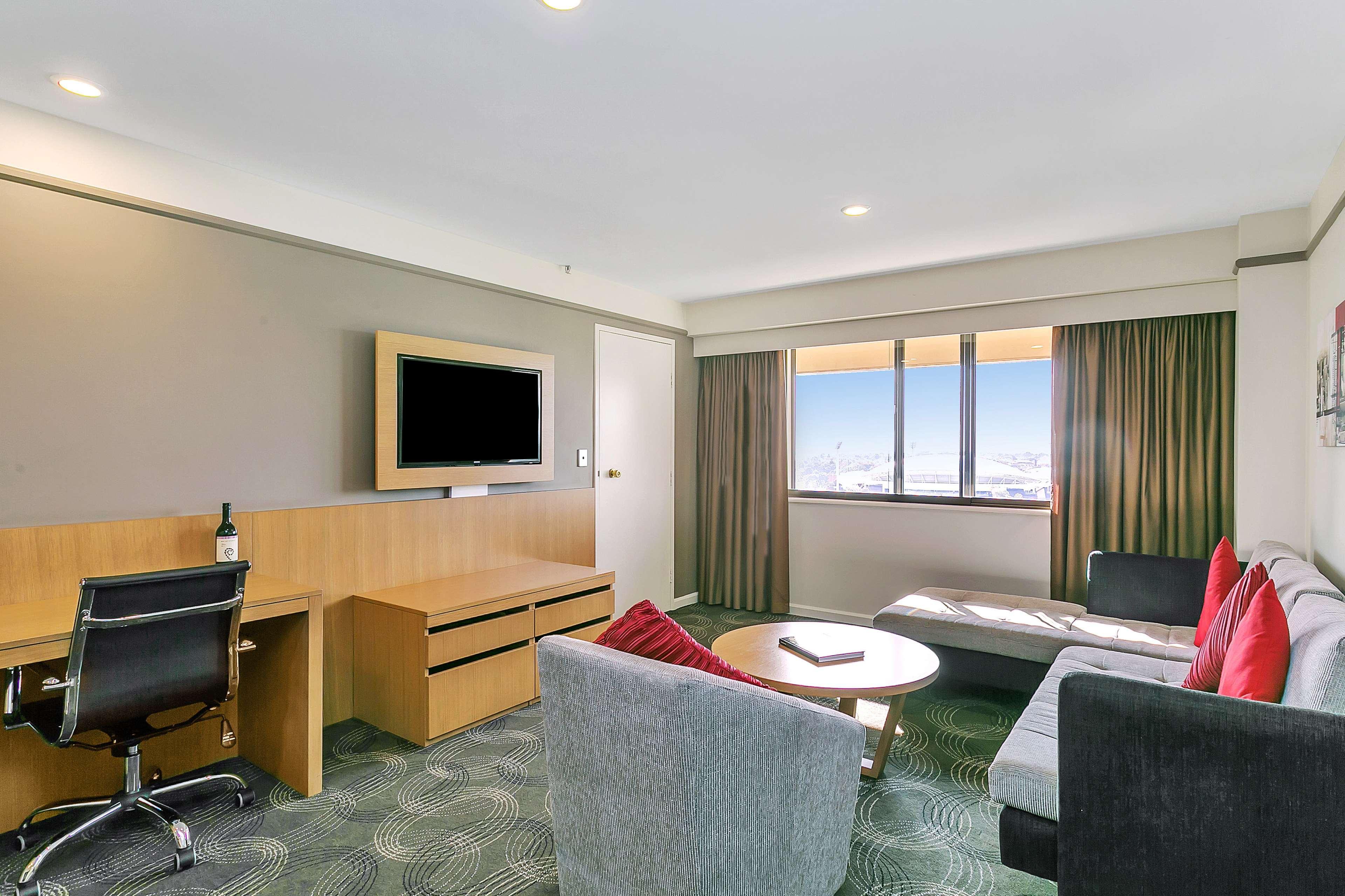 Hotel Stamford Plaza Adelajda Zewnętrze zdjęcie The photo shows a modern hotel room or suite that features a cozy seating area. There is a comfortable sofa with red accent pillows, a round coffee table in the center, and a sleek desk with an office chair. On the wall, there is a flat-screen TV mou