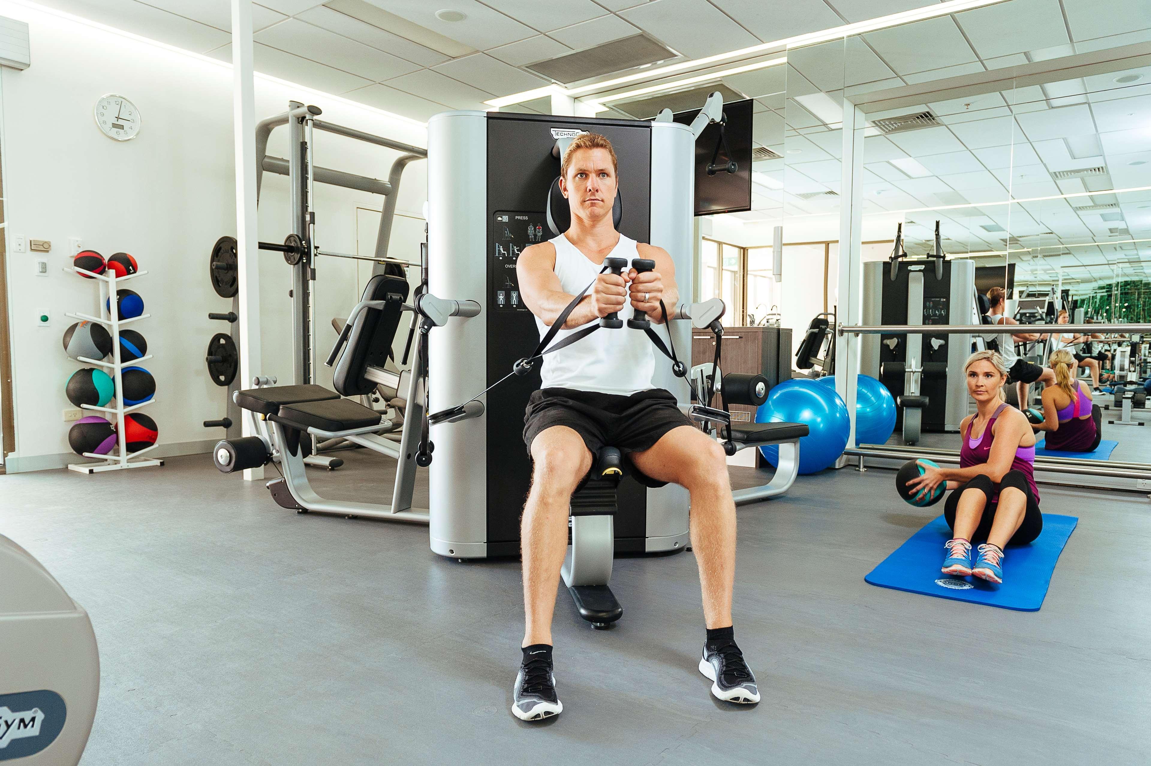 Hotel Stamford Plaza Adelajda Zewnętrze zdjęcie The photo shows a gym setting with two individuals engaged in workout activities. 

In the foreground, a man is seated on a machine, using the handles to perform a rowing exercise. He is wearing a sleeveless shirt and shorts, indicating an active wor
