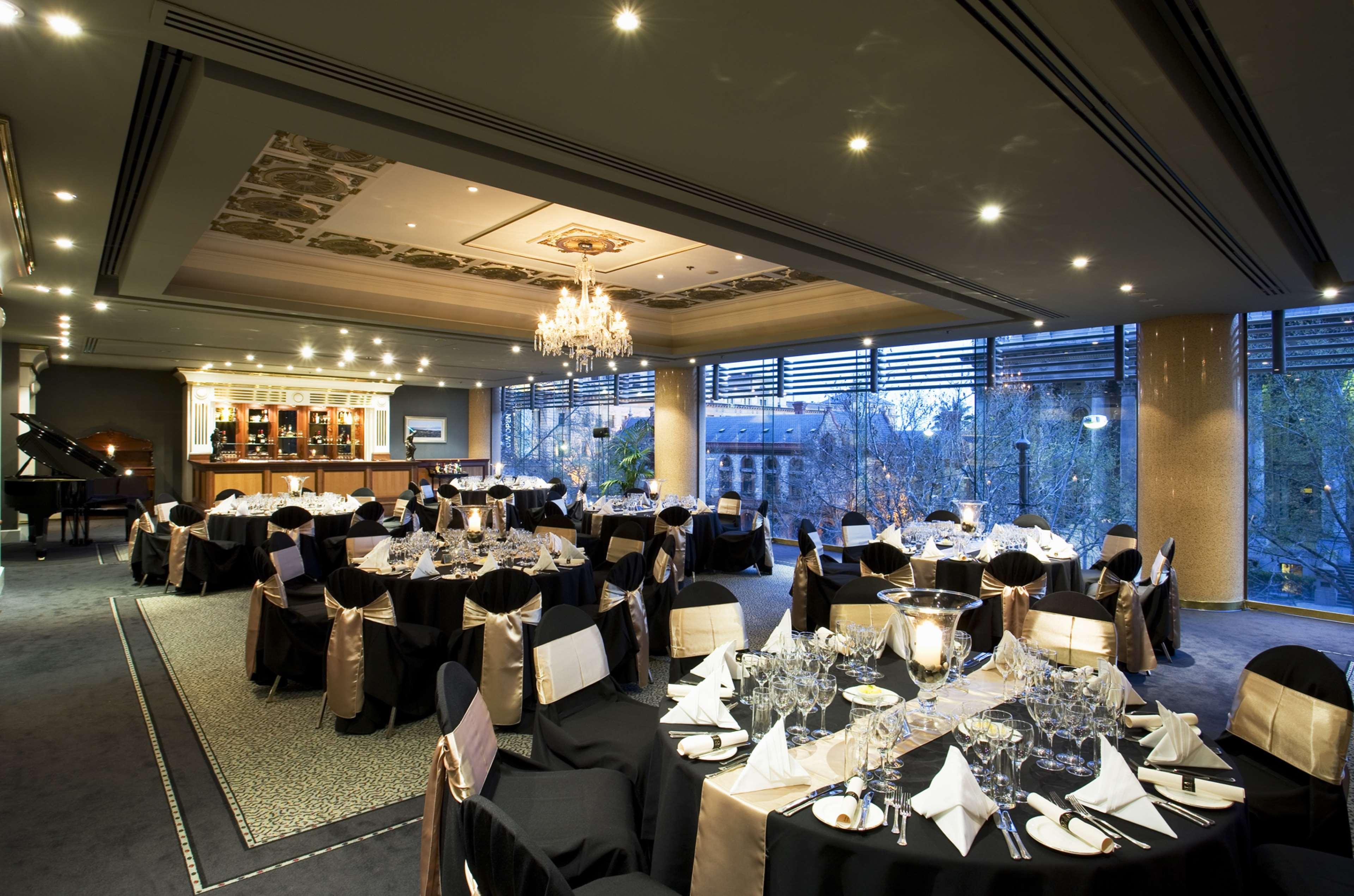 Hotel Stamford Plaza Adelajda Zewnętrze zdjęcie The photo shows an elegant banquet hall or restaurant setting. There are several round tables covered with black tablecloths and adorned with gold accents. Each table is set with fine dining utensils, glassware, and neatly folded napkins. In the back