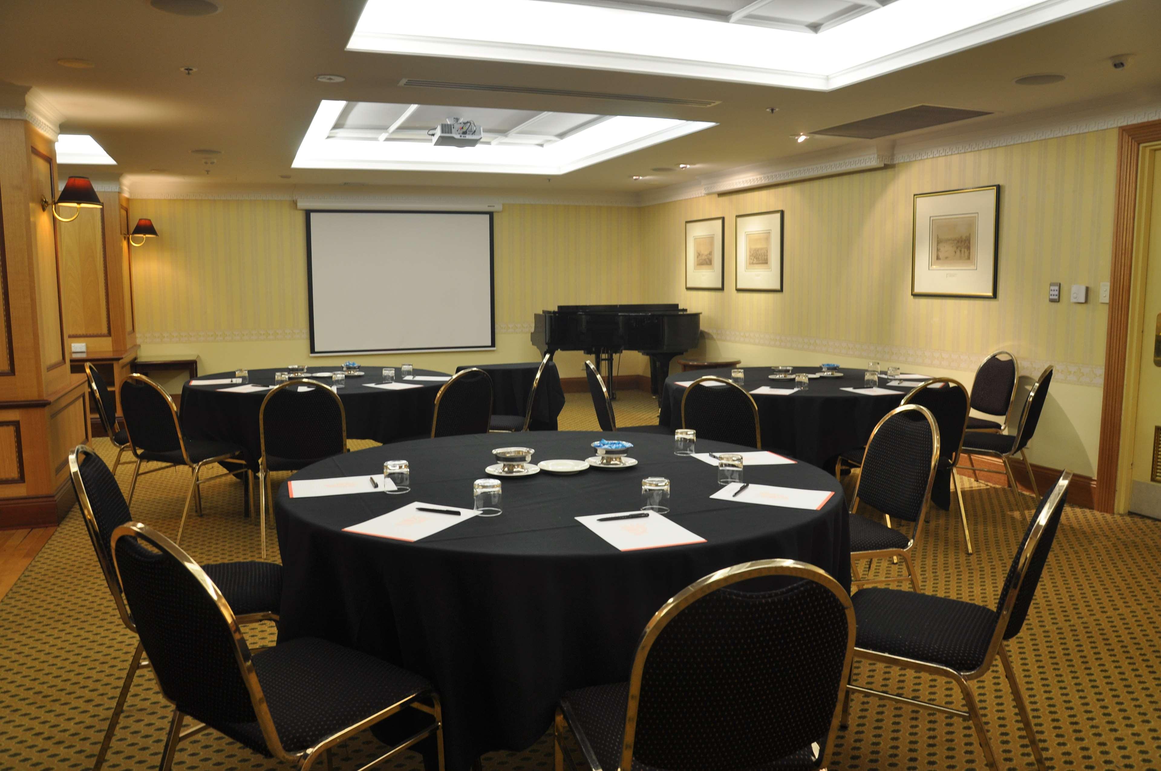 Hotel Stamford Plaza Adelajda Zewnętrze zdjęcie The photo shows a conference room set up for a meeting or presentation. There are several round tables covered with black tablecloths, each surrounded by black upholstered chairs. On the tables, there are notepads and glasses of water, indicating tha