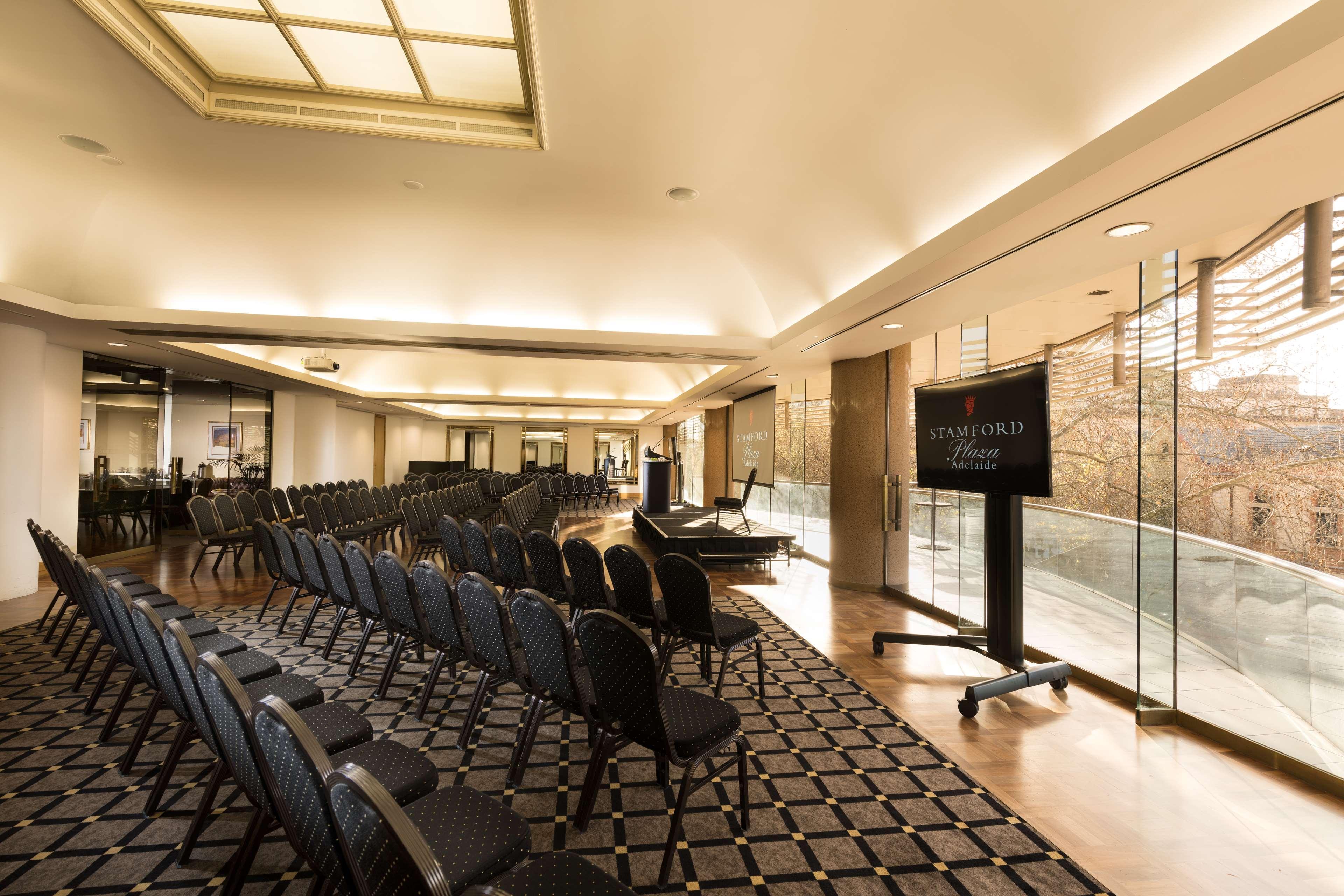 Hotel Stamford Plaza Adelajda Zewnętrze zdjęcie The photo shows a large, elegantly designed event space or meeting room. There are rows of black chairs set up facing a presentation area. The flooring features a patterned carpet, and large windows allow natural light to enter the room. On one side,
