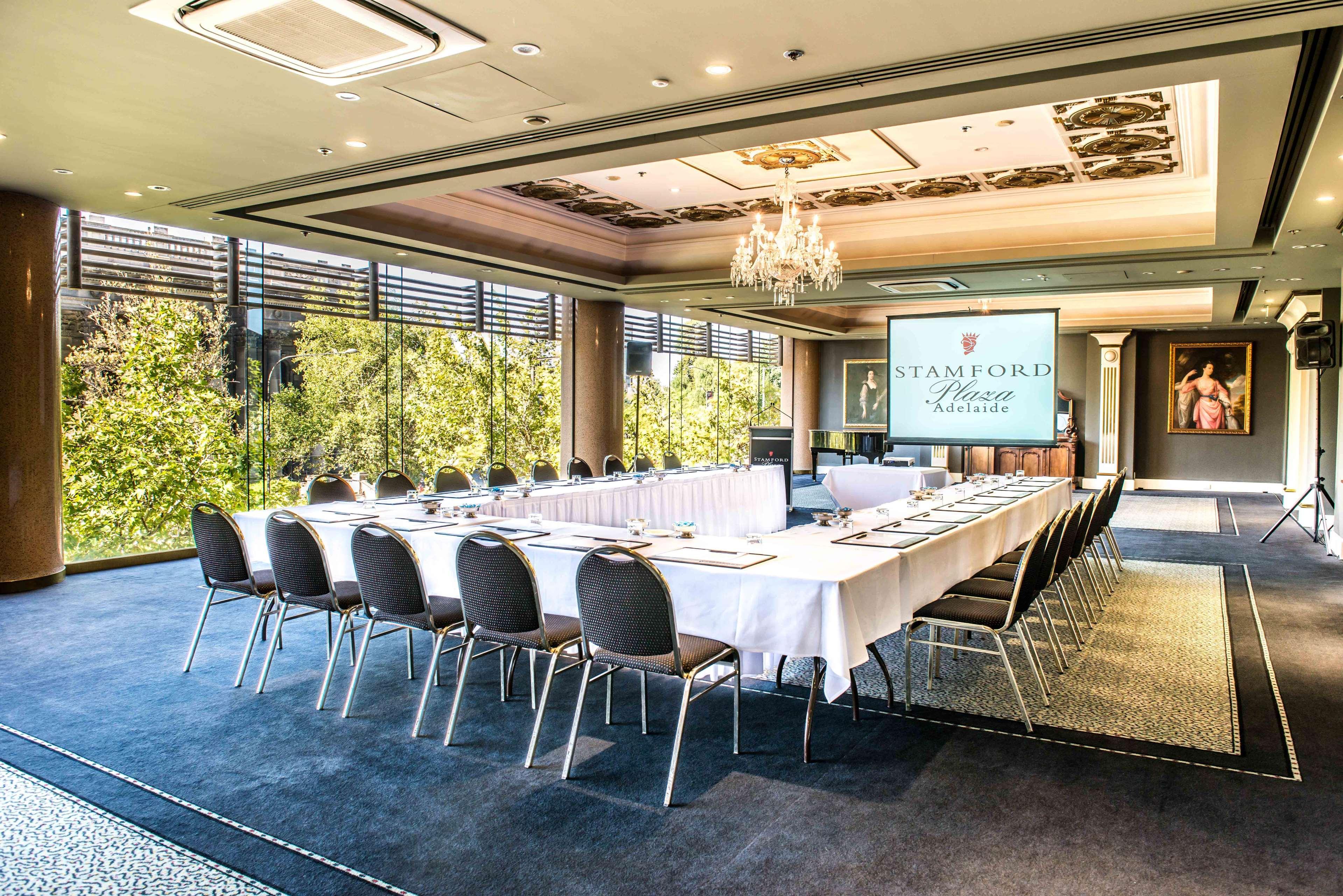 Hotel Stamford Plaza Adelajda Zewnętrze zdjęcie The photo shows a stylish conference room set up for a meeting or event. There is a long rectangular table covered with a white tablecloth, surrounded by black chairs. A large screen displays the name "Stamford" along with a logo. The room features l