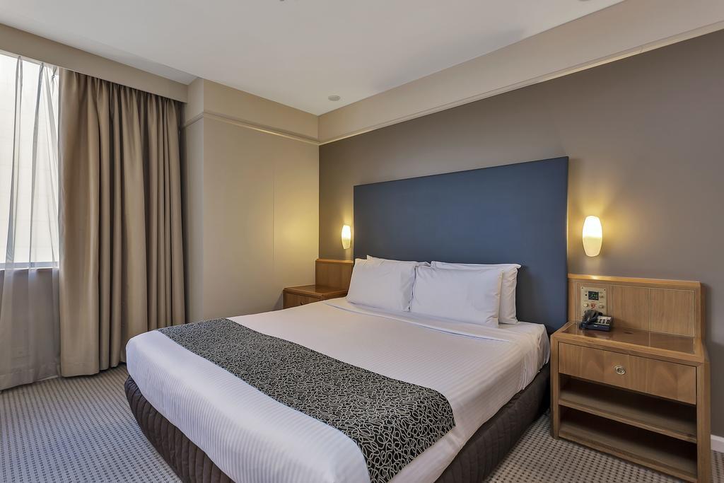 Hotel Stamford Plaza Adelajda Zewnętrze zdjęcie The photo shows a modern hotel room. It features a double bed with a white duvet and a decorative grey and black throw. The headboard is a solid dark color. There are two bedside tables on either side of the bed, each with a lamp. Natural light enter